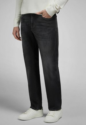 Person iført sorte jeans med lige ben, hvide sneakers og en hvid langærmet sweater med den ene hånd i lommen.