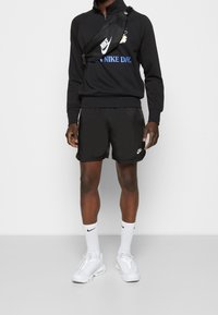 Černá mikina na zip s modrým textem a grafikou, spárovaná s černými šortkami a bílými teniskami. Na všech položkách je viditelné logo Nike.