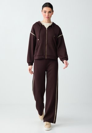 Kobieta idąca do przodu, ubrana w ciemnobrązową bluzę na zamek i pasujące szerokie spodnie z beżowym zygzakowatym wykończeniem oraz beżowe tenisówki.