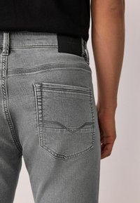 Szürke farmernadrág elegáns kialakítással, hátsó zsebek varrási részletekkel és fekete bőr címkével a deréknál.