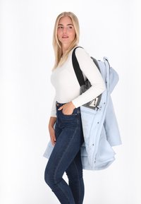Svijetloplava puffer jakna s patentnim zatvaračem i kapuljačom, sivi ruksak, bijela majica dugih rukava, tamne uske traperice, pozicija stajanja nasuprot bijeloj pozadini.