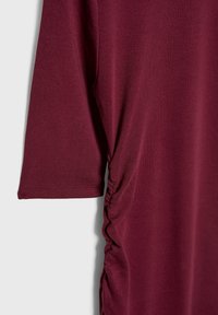 Chemise à manches courtes rouge foncé avec un détail de tissu froncé le long de la couture latérale sur un fond clair uni.