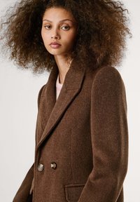 Brown double-breasted coat made of textured fabric. Notable features include large buttons and a notched collar. Model has curly hair.