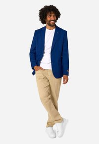 Man met krullend haar, gekleed in een blauw colbert, wit overhemd, beige broek en witte sneakers, staand met één hand in de zak.