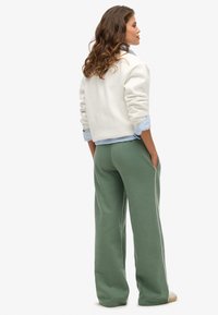 Helles graues Sweatshirt mit umschlagenen Ärmeln, über einem blauen Hemd mit Kragen getragen, kombiniert mit grünen, weit geschnittenen Jogginghosen mit Seitentaschen.