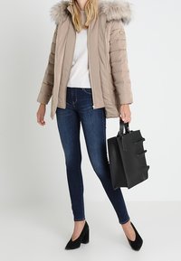 Béžová prošívaná bunda s kapucí lemovanou kožešinou, v kombinaci s tmavě modrými úzkými džíny a černými lodičkami. Černá kabelka se strukturovaným designem.