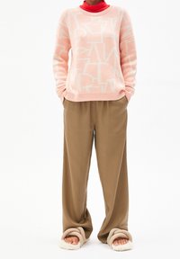 Persone che indossa un maglione a fantasia rosa chiaro, un dolcevita rosso, pantaloni marrone sciolti e pantofole beige morbide con i piedi nudi.