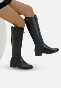 Bottes en cuir noir lisse, montantes jusqu'au genou, avec fermeture éclair sur le côté et sangle à boucle, portées par une personne assise levant une jambe.