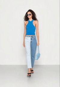 Top halter azul com nervuras, jeans com blocos de cores azul claro e escuro, sandálias pretas e uma mala azul. Os acessórios incluem óculos de sol.