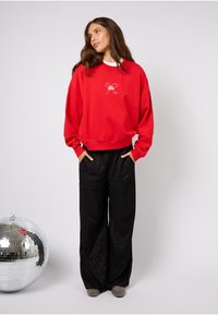 Červená mikina s grafikou bílého mašle; volný střih, žebrované manžety; doplněná černými saténovými kalhotami se širokými nohavicemi s jemným vzorem a texturovanou obuví.