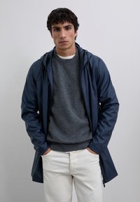 Joven con las manos en los bolsillos, vistiendo una chaqueta con capucha azul marino, un suéter gris y pantalones blancos, de pie contra un fondo claro y liso.