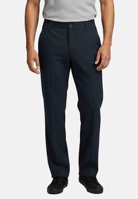 Man in zwarte rechte formele broeken, zwarte sneakers en een lichtgrijs opgestoken overhemd met korte mouwen.