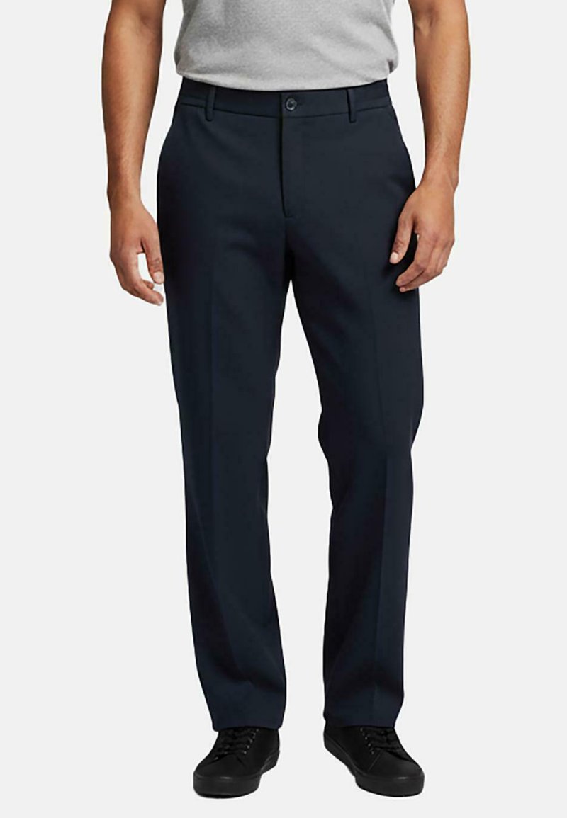 Man in zwarte rechte formele broeken, zwarte sneakers en een lichtgrijs opgestoken overhemd met korte mouwen.