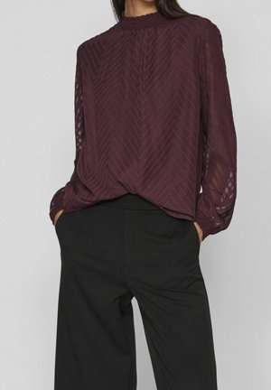 Frau trägt eine transparente langärmelige burgunderrote Bluse mit Chevronmuster, in hoch taillierte schwarze Hosen mit weitem Bein gesteckt, Hände in den Taschen.