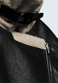 Veste en cuir noir avec un col en shearling crème, dotée d'une fermeture éclair en argent et d'une sangle à boucle noire au niveau du cou. Détails en cuir texturé visibles.