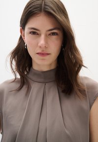 Top senza maniche in morbido tessuto beige con collo alto e pieghe frontali, caratterizzato da una superficie liscia e un design minimalista.