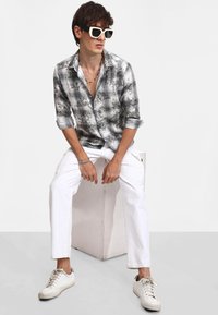 Camicia con colletto a bottoni in un motivo tie-dye nero e grigio, abbinata a pantaloni cargo bianchi e sneakers bianche. Indossati occhiali da sole angolari e di grandi dimensioni.