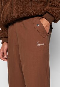 Brune, lette bukser med elastisk talje, sidelommer og et broderet hvidt logo på benet. Teksturdetaljer er synlige.