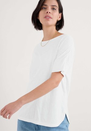 White short-sleeve shirt with a round neck, side slits, and a relaxed fit, paired with light blue denim jeans. Smooth cotton texture.