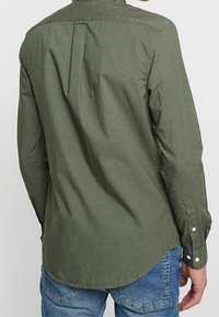 Oliwkowo-zielona koszula na guziki z plisą z tyłu, długimi rękawami i zaokrąglonym dołem. Wykonana z teksturowanej tkaniny z białymi detalami na guzikach.