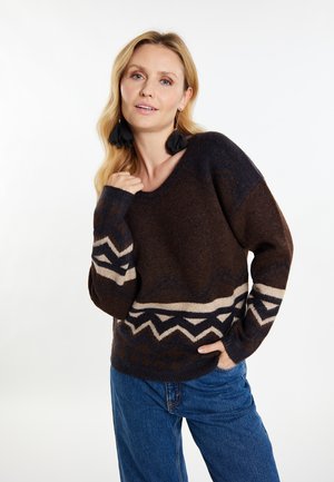 Sweter dzianinowy w ciemnym brązie z beżowymi i granatowymi wzorami geometrycznymi. Posiada dekolt w serek i długie rękawy. Nosi się go z niebieskimi dżinsami.