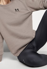 Světle hnědá mikina s černým logem "Under Armour", doplněná černými legínami s malým bílým akcentem na nohavici. Hladká textura.