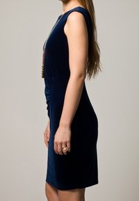 Robe en velours bleu marine avec une silhouette ajustée, des manches courtes et un détail froncé ; elle présente un collier perlé et une bague à la main gauche.