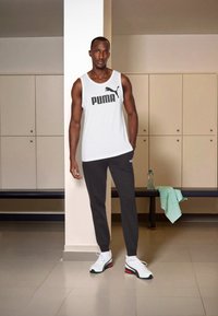 Witte mouwloze tanktop met zwart "PUMA" logo, donkergrijze broek en witte sneakers met rode en zwarte accenten. Achtergrond van een kleedkamer.