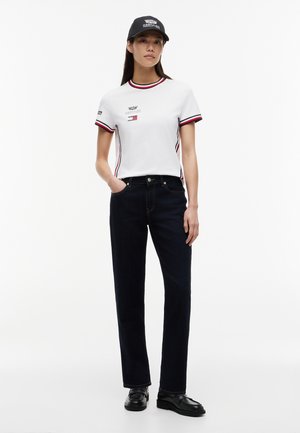 Naine, kes kannab valget keha järgivat t-särki punaste ja mustade ääristega, tumedaid teksaseid, musti loafereid ja musta baseball-mütsi, seisab valge tausta ees.