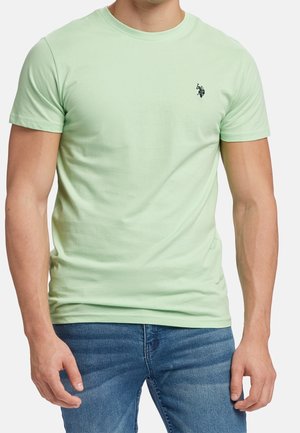 Mand iført lys grøn kortærmet t-shirt med lille sort broderet logo på venstre bryst, kombineret med blå jeans.