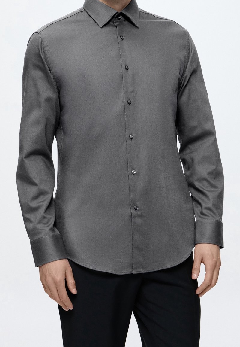 Homme portant une chemise boutonnée gris foncé à manches longues avec un pantalon noir sur un fond uni clair.