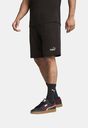 Mężczyzna ubrany w czarne szorty Puma, czarne skarpety Puma i czarne sneakersy z czerwonymi i białymi akcentami na jednolitym tle.