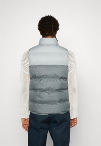 Grå puffy väst med hög krage, med en gradientdesign. Lagerindelningar skapar textur, med en slät yta och ingen synlig hårdvara.