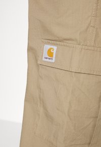 Beiga cargobyxor har ett texturerat tyg med ett rutmönster. En fyrkantig lapp med en gul logotyp är sydd på bakfickan.