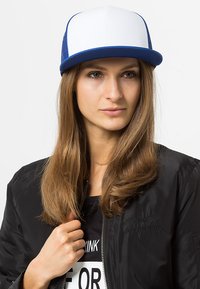 Bijela i plava trucker kapica s zakrivljenim vizirjem i mrežastim stražnjim dijelom. Nosila ju je manekenka s dugom kosom, u paru s crnom bomber jaknom.