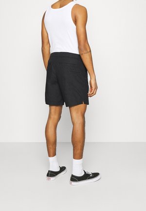 Mann trägt weißes Tanktop, schwarze Shorts, weiße Socken und schwarze Turnschuhe, steht mit dem Rücken zur Kamera vor einem einfarbigen Hintergrund.