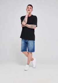 Čierne tričko s krátkym rukávom, modré džínsové šortky a biele tenisky. Tetovania viditeľné na ramenách; voľný strih a jednoduchý dizajn.