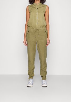 Femme portant une combinaison sans manches vert olive avec boutons à l'avant, taille ajustable par cordon, poches zippées et baskets blanches, debout contre un mur blanc.