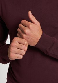 Pull bourgogne avec des poignets côtelés, gros plan sur des mains ajustant le tissu. Texture lisse et design ajusté.