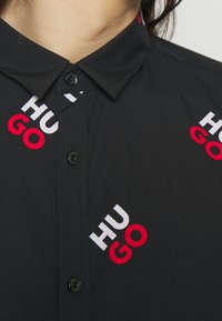 Czarna koszula na guziki z kołnierzykiem, ozdobiona rozproszonym napisem "HUGO" w kolorach czerwonym i białym. Gładka tkanina z delikatnym połyskiem.