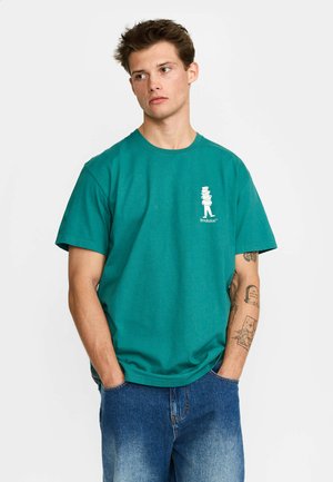 Joven con tatuajes que lleva una camiseta verde azulado con un pequeño gráfico blanco y vaqueros azules holgados, con las manos en los bolsillos, mirando hacia un lado.