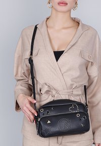 Vrouw in beige blazer met strik, die een zwarte leren schoudertas vasthoudt, versierd met studs in de vorm van sterren, een hart en een cirkel met een "N" erin.