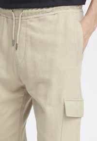 Beige sweatpants met een elastische tailleband en trekkoord. Voorzien van zijzakken en een cargozak op de dij. Zachte stofstructuur.
