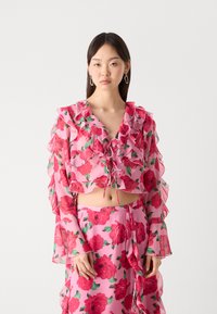 Blusa cropped floreale rosa con maniche a volant e scollatura con lacci. Presenta rose vivaci e tessuto fluido con una consistenza leggera.