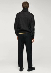 Schwarzer gestrickter Rollkragenpullover mit einem strukturierten Muster, kombiniert mit schwarzen Hosen und schwarzen Schuhen. Der Hintergrund ist minimalistisch und neutral.