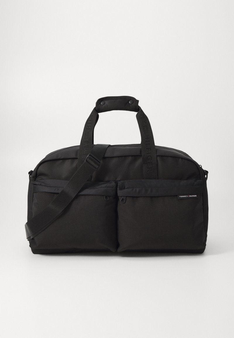 Black fabric duffel bag with dual front pockets, padded handles, adjustable shoulder strap, and logo detailing on the strap.