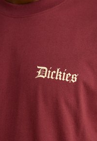 T-shirt en coton bordeaux avec un logo brodé couleur crème "Dickies" en police gothique sur la poitrine gauche. Design classique à col rond.