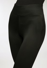 Czarne legginsy z wysokim stanem noszone przez osobę, ukazujące gładki, dopasowany materiał od talii do górnej części ud na białym tle.