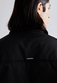 Homme aux cheveux foncés mi-longs, portant une veste noire avec le logo "SANDRO" en haut du dos, dos à la caméra.