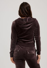 Felpa in velluto marrone con una texture morbida, dotata di cappuccio, e pantaloni della tuta abbinati con due tasche posteriori e dettagli a bottoni a pressione.